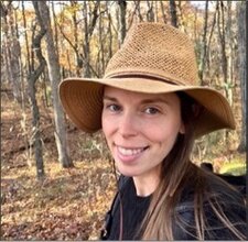 A young woman wearing a broad brimmed hat is smiling for the camera in an autumn woodland scene.