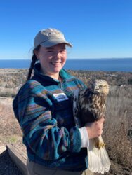 Young woman smiling, wearing a ball cap and holding an owl. The background is open ground with water on the horizon and clear blue skies.