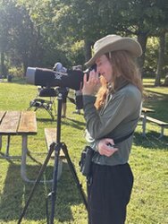 A side view of a woman looking into a spotting scope on a tripod. The foreground is open with picnic tables. A line of trees forms the background.