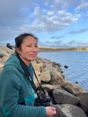 A young woman sitting on a rocky shore looking to her right. A camera is around her neck. A shoreline runs from middle right to the horizon behind her. A line on land separates the water from the partly sunny sky.